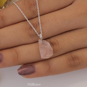 Puede incluir: Un collar con un colgante de cuarzo rosa en bruto de color rosa pálido suspendido de una fina cadena de metal plateado. La piedra tiene una forma irregular y facetada con un aspecto translúcido. La palabra Sillure está impresa en la imagen.