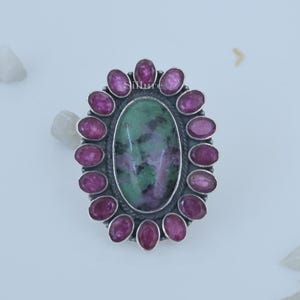 Op de afbeelding: Een zilveren ring met een grote ovale groene en roze edelsteen, omringd door een halo van kleinere roze edelstenen.