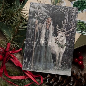 May include: A rectangular art print featuring an elderly person with long white hair wearing a dark hooded cloak and holding a staff, standing beside a white reindeer with a wreath around its neck. The text reads mary feywood ART BY MAARTJE VAN DORKUM.