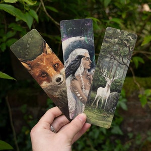 May include: Three rectangular bookmarks with fantasy art. One features a fox with the word "feywood". Another depicts a woman with a raven, and the third shows a white deer in a forest. The bookmarks are held against a backdrop of green foliage.