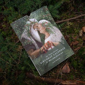 Könnte beinhalten: Ein Spiral-Kalender mit einer Illustration von zwei Figuren mit Blumenkränzen, die sich umarmen. Der Kalender mit dem Titel "BIRTHDAY CALENDAR" befindet sich vor einem Hintergrund aus immergrünem Laub und Moos. Der Text "the forest witches" und "With monthly full moon names" ist ebenfalls sichtbar.