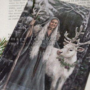 May include: A rectangular art print featuring a painting of an elderly person with long white hair wearing a dark hooded cloak and holding a staff, standing next to a white reindeer with antlers and a green wreath. Text reads mary feywood.