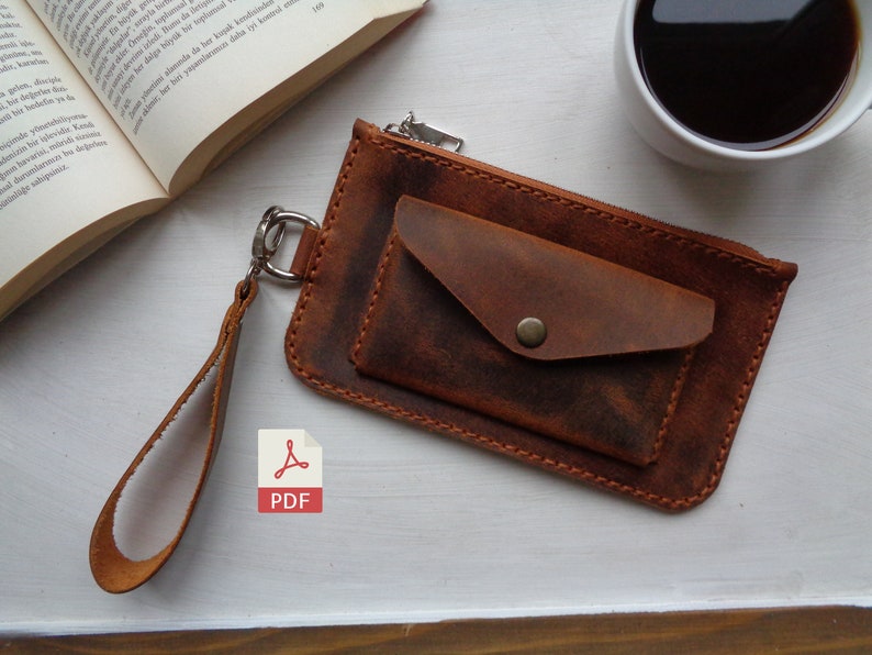 May include: A brown leather wallet with a zipper closure and a small pocket on the front. The wallet has a brown leather strap with a metal ring.