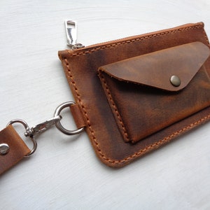 May include: A brown leather wallet with a zipper closure and a flap pocket. The wallet has a strap with a metal clip.