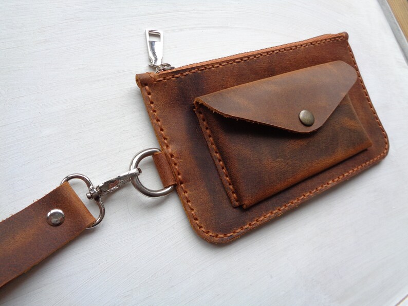 May include: A brown leather wallet with a zipper closure and a flap pocket. The wallet has a strap with a metal clip.