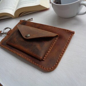May include: A brown leather pouch with a zipper closure and a smaller flap pocket. The pouch has a strap with a metal ring.