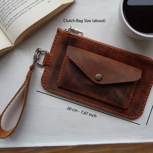 May include: A brown leather clutch bag with a zipper closure and a small pocket on the front. The bag has a wrist strap and measures 7.87 inches by 4.33 inches.