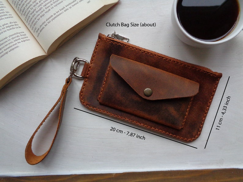 May include: A brown leather clutch bag with a zipper closure and a small pocket on the front. The bag has a wrist strap and measures 7.87 inches by 4.33 inches.