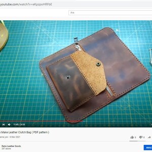 May include: A brown leather clutch bag with a zipper closure and a smaller pocket on the front. The bag is lying on a green cutting mat.