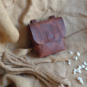 Peut inclure: Un sac en cuir brun avec une fermeture &agrave; rabat et un petit patch carr&eacute; sur le devant. Le sac est pos&eacute; sur un fond en toile de jute.