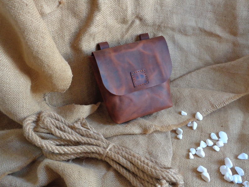 Peut inclure: Un sac en cuir brun avec une fermeture &agrave; rabat et un petit patch carr&eacute; sur le devant. Le sac est pos&eacute; sur un fond en toile de jute.