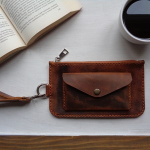May include: A brown leather wallet with a zipper closure and a small pocket on the front. The wallet has a wrist strap and a silver zipper pull.