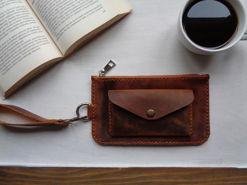 May include: A brown leather wallet with a zipper closure and a small pocket on the front. The wallet has a wrist strap and a silver zipper pull.