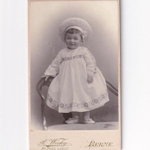 Könnte beinhalten: Vintage Schwarz-Weiß-Fotografie eines kleinen Kindes in einem weißen Kleid und Hut. Das Kleid hat gestickte Details, und das Kind steht in der Nähe eines Stuhls. Die Fotografie ist signiert "A. Wicky, Fr. Fuss sucer" und "BERNE."