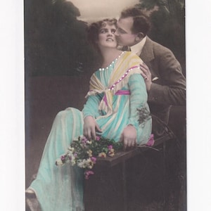 May include: A vintage sepia postcard showing a couple embracing. The woman is in a long, light blue dress with a striped shawl and flowers. The man kisses her cheek. The background is a blurred outdoor scene.