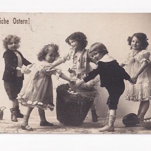 Può includere: Cartolina vintage in seppia con la scritta "Fröhliche Ostern!" in alto. Cinque bambini sono riuniti attorno a un grande cesto pieno di uova e una gallina. I bambini si tengono per mano, festeggiando la Pasqua.