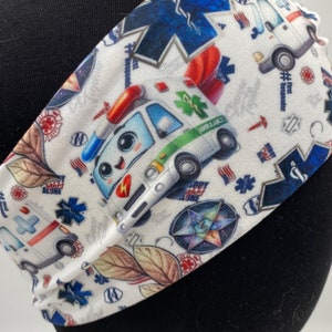 May include: White headband with a colorful pattern of emergency vehicles, stars, and medical symbols. The pattern includes a cartoon ambulance, a cartoon police car, and a red heart with a bandage. The text "#FirstResponder" is visible on the headband.