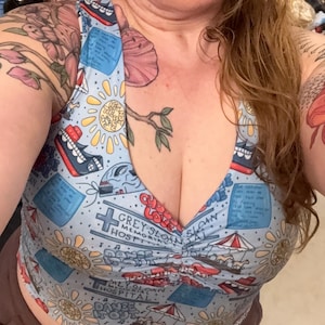 May include: A light blue sleeveless crop top featuring a repeating pattern of Grey Sloan Memorial Hospital logos, yellow suns, red and white carousels, blue sticky notes, and red hearts with the text You're My Person and Dance It Out.