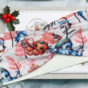 May include: A white headband with a pink, blue and red Christmas-themed print featuring an octopus, a penguin and a dolphin. The print also includes a Christmas tree and snowflakes.