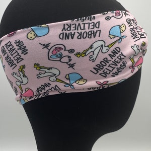 May include: Pink headband with a repeating pattern of storks delivering babies, hearts, and the text "Labor and Delivery Nurse".