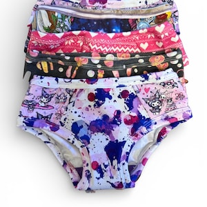 May include: A stack of colorful underwear with various patterns. The designs include a purple and blue splatter print, hearts, and cartoon characters. The elastic waistbands are visible, and the fabric appears soft.