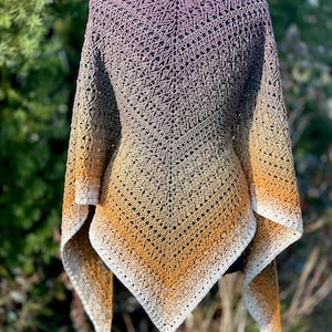 May include: A handcrafted, open-weave shawl in a gradient of colours, transitioning from purple to gold. The shawl is displayed on a mannequin, showcasing its intricate crochet pattern and flowing design. The edges are finished with a white border.