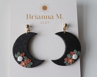 Puede incluir: Pendientes de media luna negra con purpurina y detalles florales en blanco, rosa y verde. Brianna M. CLAY.