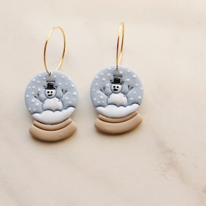 May include: A pair of whimsical snowman snow globe earrings. Each earring features a light blue globe with a snowman, white snow, and a tan base. The earrings hang from gold-colored hoops.