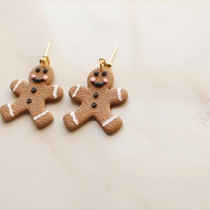 May include: A pair of gingerbread man earrings with gold hooks. The gingerbread men are brown with white icing and black buttons and eyes. They have pink cheeks and a smiling face.