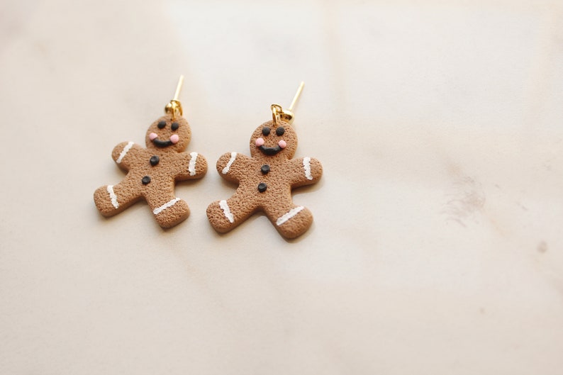 May include: A pair of gingerbread man earrings with gold hooks. The gingerbread men are brown with white icing and black buttons and eyes. They have pink cheeks and a smiling face.