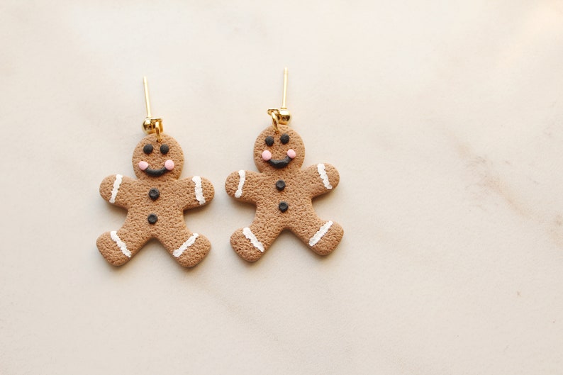 May include: A pair of gingerbread man earrings with white icing and black eyes. The earrings are made of polymer clay and have gold hooks.