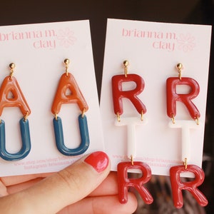 May include: Two pairs of polymer clay earrings. The first pair features the letters "AU" in orange and blue. The second pair features the letters "RR" in red and white. The earrings are on a white card with the text "brianna m. clay" printed on it.
