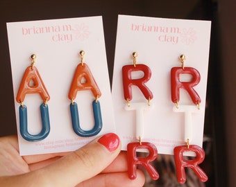 May include: Two pairs of polymer clay earrings. The first pair features the letters "AU" in orange and blue. The second pair features the letters "RR" in red and white. The earrings are on a white card with the text "brianna m. clay" printed on it.