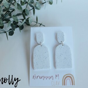 May include: White speckled clay earrings with a rounded arch shape. The earrings are on a white card with the text "Briana M Clay" and a rainbow.