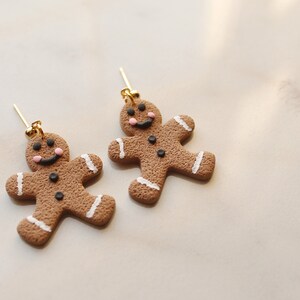 May include: A pair of gingerbread man earrings made of brown clay with white icing and black details. The earrings have gold hooks.
