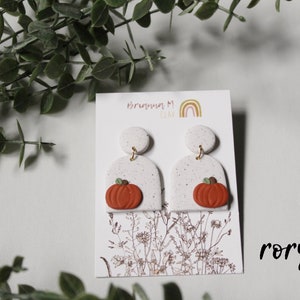 May include: White and orange pumpkin dangle earrings with gold accents. The earrings are on a white card with the text "Brenna M. Clay" and "Rory".