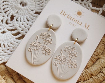 May include: A pair of off-white clay earrings. Each earring features a circular top and an oval pendant with a daisy design. The earrings are connected with gold-colored hoops. The brand name "Brianna M. Clay" is printed on the card.