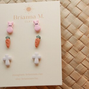 May include: A set of six pastel-colored clay earrings featuring a pink bunny, a carrot, and a white cross. The earrings are displayed on a white card with the text "Brianna M. Clay" printed on it.