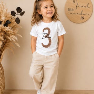 Könnte beinhalten: Ein weißes Kurzarm-T-Shirt mit einer großen braunen Glitzer-Zahl Drei und dem Namen Ida in schwarzer Schreibschrift in der Mitte. Das Oberteil wird mit einer beigen Hose und weißen Turnschuhen kombiniert, ergänzt durch eine kleine Haarspange.