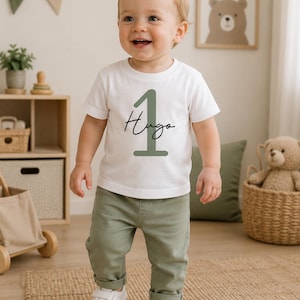 Könnte beinhalten: Ein weißes kurzärmeliges T-Shirt mit einer großen salbeigrünen Zahl Eins und dem Namen Hugo in schwarzer Schreibschrift in der Mitte, kombiniert mit einer salbeigrünen Hose und weißen Turnschuhen mit Klettverschluss.