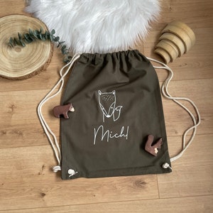 Puede incluir: Una bolsa de cord&oacute;n verde con una ilustraci&oacute;n de zorro blanco y el nombre "Michl" impreso en ella. La bolsa est&aacute; hecha de lona y tiene cordones de cord&oacute;n blancos.