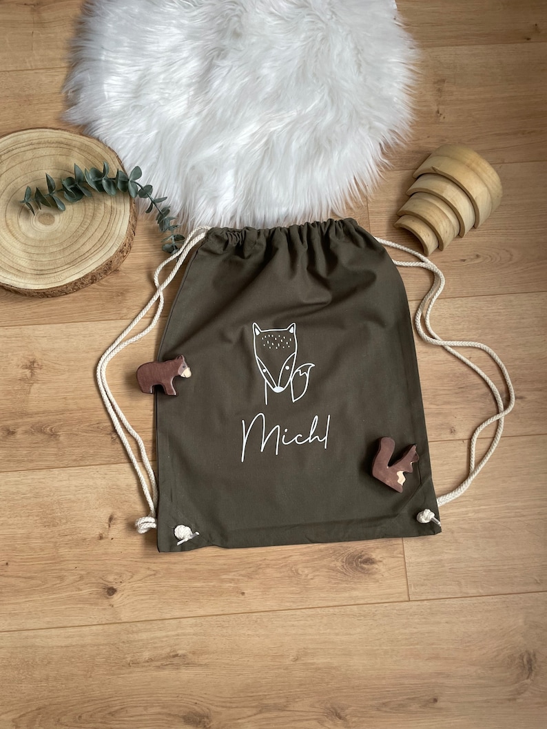 Puede incluir: Una bolsa de cord&oacute;n verde con una ilustraci&oacute;n de zorro blanco y el nombre "Michl" impreso en ella. La bolsa est&aacute; hecha de lona y tiene cordones de cord&oacute;n blancos.