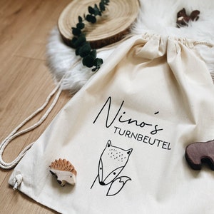 Puede incluir: Una bolsa de cord&oacute;n blanco con el texto "Nino's Turnbeutel" y un dibujo de l&iacute;nea negra de un zorro. La bolsa est&aacute; sobre una superficie blanca y esponjosa con figuritas de animales de madera.