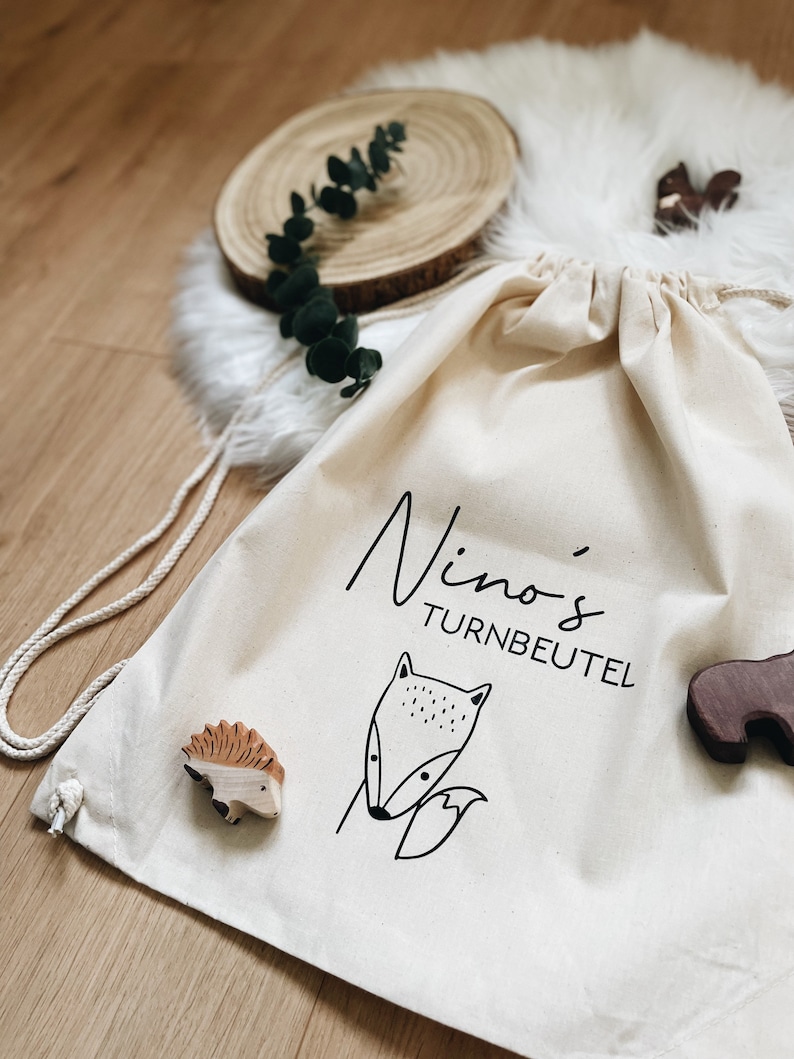 Puede incluir: Una bolsa de cord&oacute;n blanco con el texto "Nino's Turnbeutel" y un dibujo de l&iacute;nea negra de un zorro. La bolsa est&aacute; sobre una superficie blanca y esponjosa con figuritas de animales de madera.