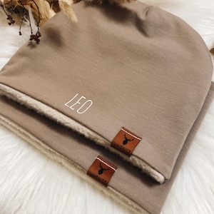 Könnte beinhalten: Eine braune Mütze mit weißem Fleecefutter. Die Mütze hat den Namen "LEO" auf der Vorderseite gestickt und ein braunes Leder-Patch mit einem Hirsch-Logo und dem Text "WAIDKNITZ" an der Seite.