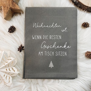Könnte beinhalten: Ein gefaltetes graues Textil mit weißer Schrift, die lautet: "Weihnachten ist... WENN DIE BESTEN GESCHENKE AM TISCH SITZEN" und einem kleinen silbernen Glitzertannenbaum. Das Textil ist von dekorativen Gegenständen auf einer weißen, flauschigen Oberfläche umgeben.