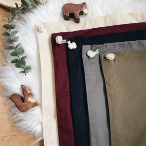 Puede incluir: Un conjunto de seis cuadrados de tela en varios colores, incluyendo beige, burdeos, negro, gris y verde oliva. Cada cuadrado tiene un detalle de cuerda anudada en la esquina.