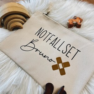Könnte beinhalten: Eine beige Canvas-Reißverschlusstasche mit den Worten "NOTFALLSET Bruno" und einer braunen Pflastergrafik. Holzspielzeug, darunter ein Eichhörnchen und ein Pferd, sind um die Tasche auf einer weißen, flauschigen Oberfläche verstreut.