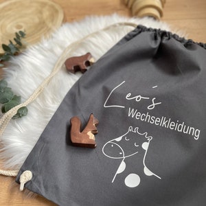 Puede incluir: Bolsa de cord&oacute;n gris con texto blanco que dice "Leo's Wechselkleidung" y una ilustraci&oacute;n blanca de una jirafa con manchas blancas. La bolsa est&aacute; sobre una superficie blanca y esponjosa.