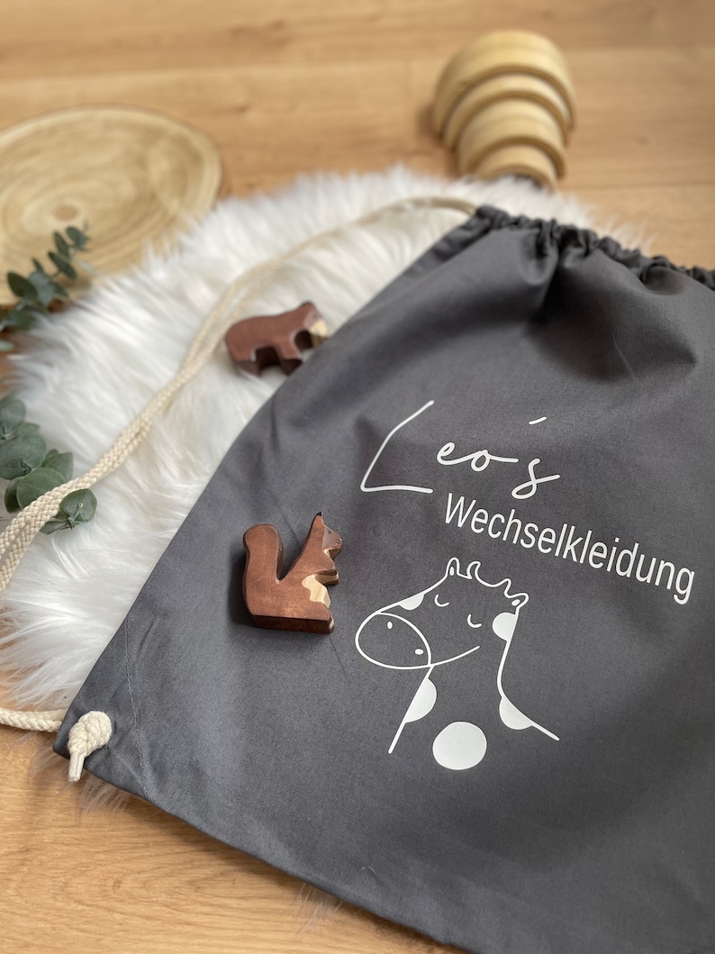 Puede incluir: Bolsa de cord&oacute;n gris con texto blanco que dice "Leo's Wechselkleidung" y una ilustraci&oacute;n blanca de una jirafa con manchas blancas. La bolsa est&aacute; sobre una superficie blanca y esponjosa.